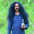 Man with long dark hair holding a hakki pikki hair growth oil bottle outdoors